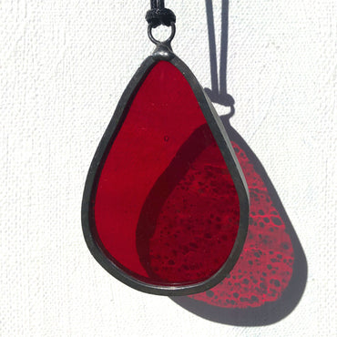a small red blood drop stained glass charm with translucent red glass against a white wall, red glass refractions shine onto the white background